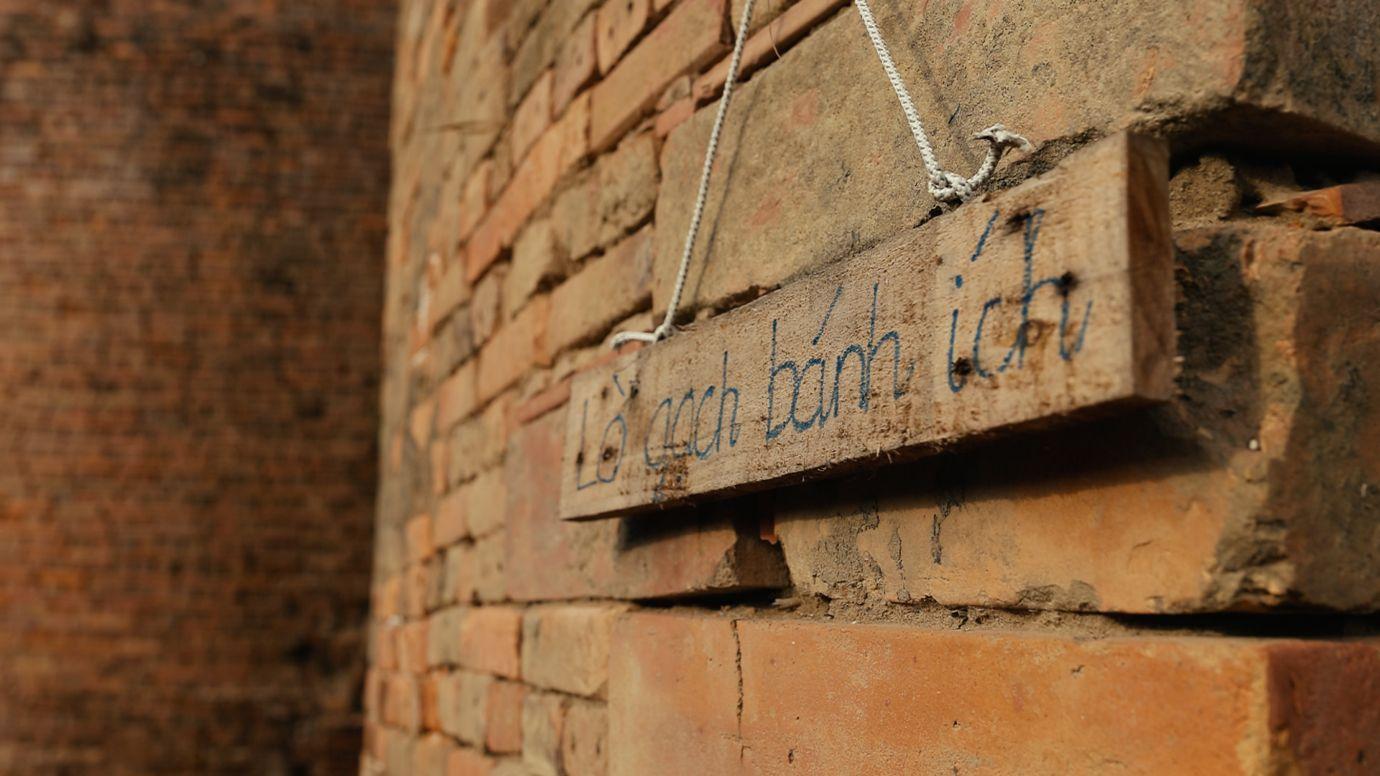 A rustic wooden signboard reading 'bánh ít brick kiln' hung on a moss-covered wall, an interesting highlight when visitors come to the Hoa Sen brick-kiln homestay.