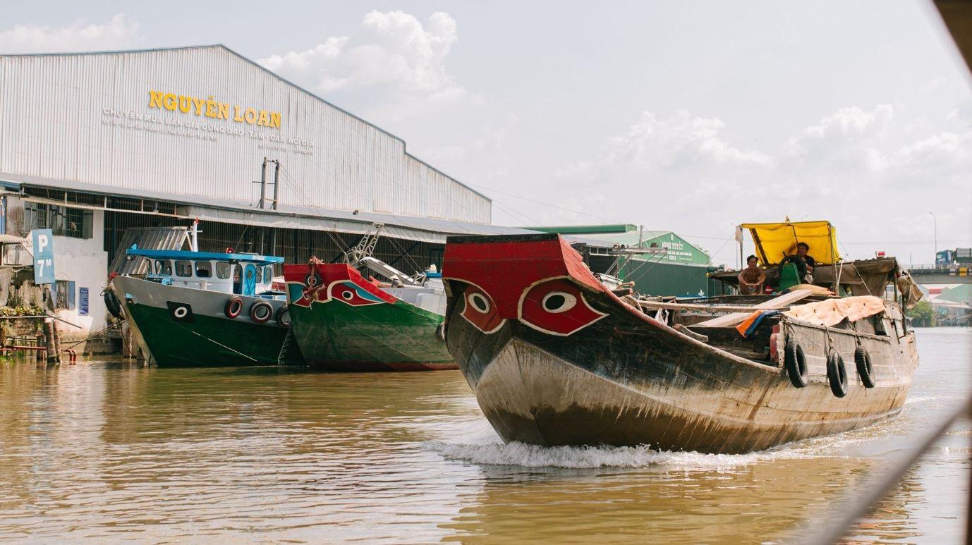 A wooden boat with traditional 'boat-eye' motifs cutting through the waves past large rice mills, carrying the rhythm and soul of Sa Đéc's merchant-river culture across generations.